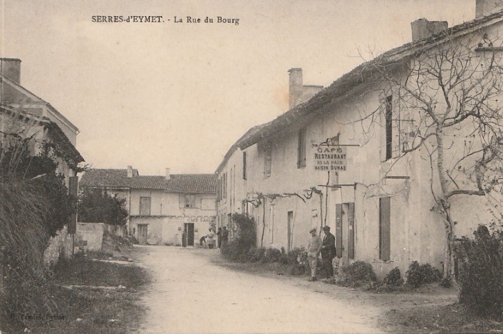 Old black-and-white street scene in Serres-d'Eymet, La Rue du Bourg, showing white village houses, a dirt road, leafless trees, and a café/restaurant sign with two people standing nearby.